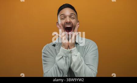 Giovane uomo che esprime gioia su un vivace sfondo arancione indossando una camicia grigia casual e sorridendo ampiamente e coinvolgendo gli spettatori con energia e unicità Foto Stock