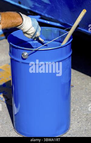 Secchio in metallo con vernice blu. Una mano in un guanto protettivo tiene in mano una spazzola. Verniciatura di prodotti metallici Foto Stock