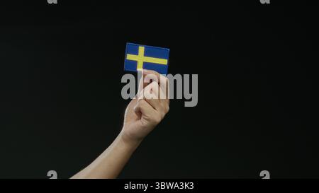 Mano che regge una bandiera svedese su sfondo nero, simboleggiando la nazionalità e l'orgoglio svedesi, isolata e prominente nella scena, capturin Foto Stock