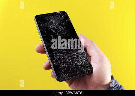Smartphone nero con touch screen rotto in mano su sfondo giallo. Nella mano di un uomo c'è un dispositivo mobile con vetro incrinato Foto Stock