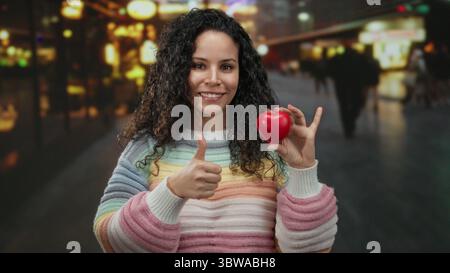 Giovane donna ispanica che tiene in mano un oggetto rosso a forma di cuore e i pollici gestanti in strada illuminata con sfondo sfocato sotto l'illuminazione notturna. Foto Stock