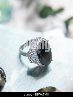 Anello in argento con pietre preziose nere e diamanti su sfondo azzurro con foglie verdi sfocate Foto Stock
