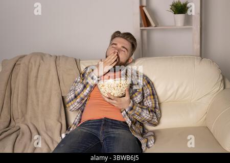 Uomo barbuto che guarda film o giochi sportivi in TV mangiando popcorn in casa la sera. Cinema, campionato e fan concept Foto Stock