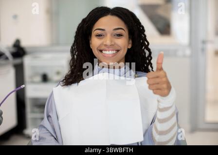 Sorridente femmina nera che sorride e mostra il pollice su dopo il trattamento dentale Foto Stock
