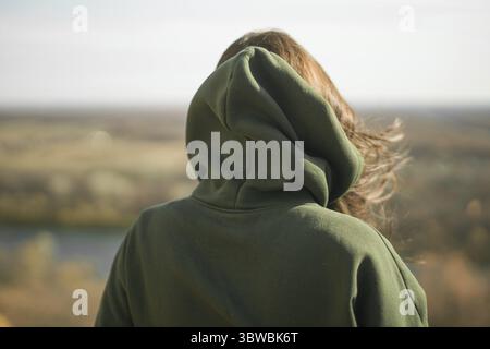 Vista posteriore di una giovane donna alla moda con una felpa con cappuccio verde scuro in autunno. Un simpatico modello cammina nel parco in autunno dorato tra alberi colorati e foglie cadute. Foto Stock