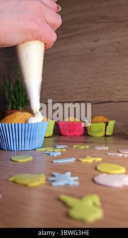 Versare a mano la panna su un cupcake in una cassa blu con custodie colorate sullo sfondo. Spruzzi decorativi sparsi su un tavolo di legno Foto Stock