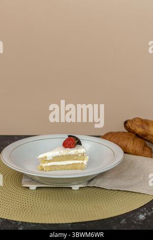 Una fetta di deliziosa torta a strati con glassa di panna bianca, condita con una fragola fresca e un biscotto. Un dessert perfetto con croissant sul retro. Foto Stock