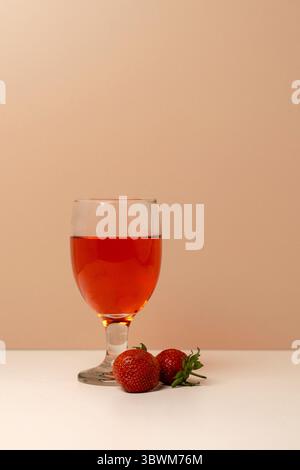 Natura morta minimalista di un bicchiere di succo di fragola fresco o di vino rosa con due fragole mature. Ampio spazio di copia su sfondo beige. Foto Stock