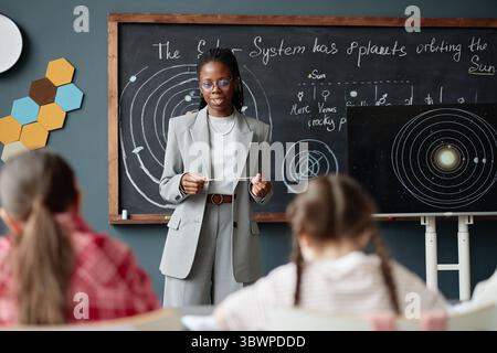 Insegnante di donna nera che spiega il diagramma del sistema solare a un gruppo di bambini in classe, tenendo il puntatore e in piedi di fronte alla lavagna con disegni planetari, gli studenti ascoltano attentamente Foto Stock