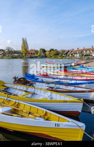 Colorate barche a remi ormeggiate a Thorpeness Meare nel Suffolk, Inghilterra. Sullo sfondo, i cigni nuotano sull'acqua. Case sulla riva opposta. Foto Stock