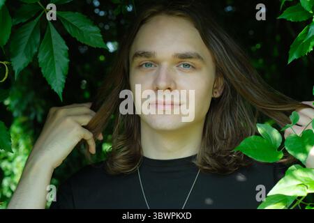 Un giovane sorride tenendo delicatamente i suoi lunghi capelli in un ambiente verde vivace. Foto Stock