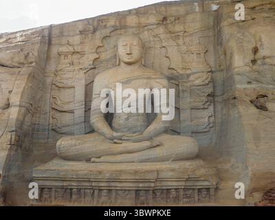 Antica statua scolpita nella roccia di un Buddha seduto in posizione di meditazione, circondata da sculture in pietra, situata nello Sri Lanka. Foto Stock