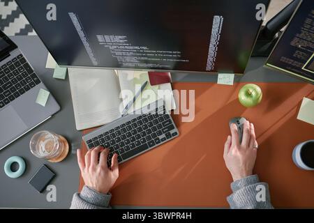 Giovane adulto caucasico che lavora alla scrivania del computer utilizzando tastiera e mouse, scrivendo codice sul monitor, circondato da forniture per ufficio e dispositivi elettronici, area di lavoro con vista dall'alto Foto Stock