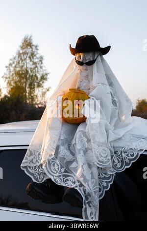 il bambino con coperta bianca, cappello, occhiali neri con una zucca arancione scolpita per le vacanze siede sul tetto di un'auto bianca. Persone in costumi fantasma per Halloween Foto Stock