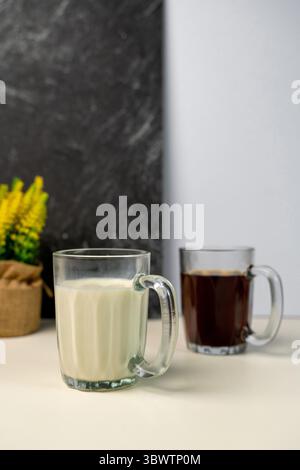 Bevanda mattutina a scelta: Tazza di vetro con latte fresco o caffè nero. Fiori gialli di Solidago sullo sfondo, su una moderna parete testurizzata. Foto Stock