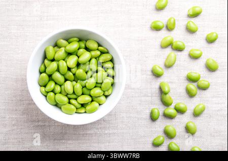 Fagioli di edamame in una ciotola bianca su lino. Soia giovane e immatura sgusciata, Glycine max, di colore verde, spesso mangiata come spuntino, al vapore o bollita. Foto Stock