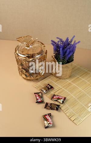 Giacarta, Indonesia, 17 luglio 2025: Vaso decorativo di caramelle in vetro ambrato con coperchio a farfalla, caramelle per caffè e lavanda su un tappetino in bambù. Foto Stock
