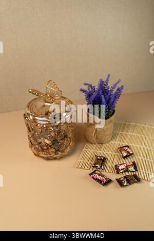 Giacarta, Indonesia, 17 luglio 2025: Vaso decorativo di caramelle in vetro ambrato con coperchio a farfalla, caramelle per caffè e lavanda su un tappetino in bambù. Foto Stock