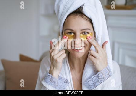 Tempo libero a casa, trattamenti spa e cura delicata della pelle. Rilassatevi e distendetevi. Bella donna caucasica in asciugamano applica patch sotto gli occhi seduti in l Foto Stock