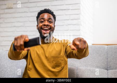 Eccitato giovane afroamericano che guarda lo sport in tv, festeggia il successo e urla mentre siede sul divano a casa sua. Foto Stock