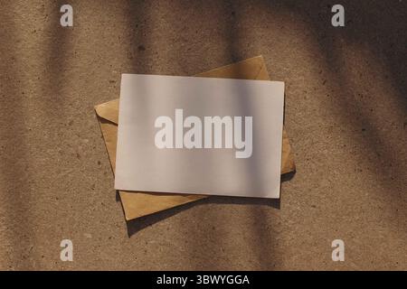 Mockup di invito per biglietti d'auguri vuoti orizzontali minimalisti, busta di carta artigianale. Sfondo in pietra neutro. Ombre floreali morbide e naturali, calde Foto Stock