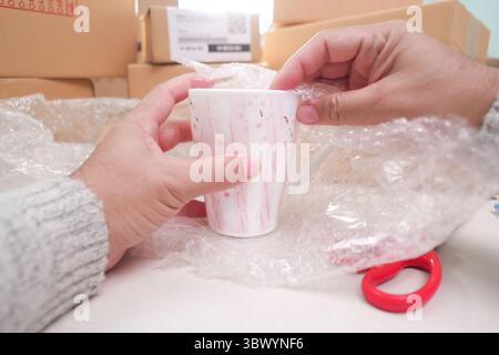 Mani che disimballano o confezionano una tazza in ceramica con pellicola a bolle d'aria, che mostra la cura degli articoli fragili, la spedizione, circondata da altri pacchi di consegna. Foto Stock