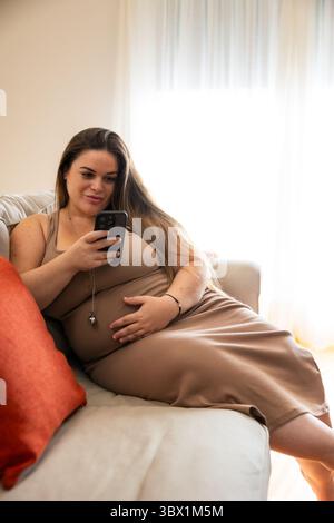 Aspettati una donna in un abito beige comodamente seduta su un divano, tenendo la pancia con una mano e sfogliando il suo smartphone con un'expres calma e mirata Foto Stock