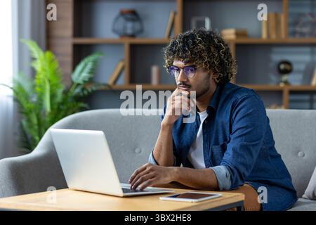 Un giovane uomo con capelli ricci e occhiali lavora su un laptop, nel profondo del pensiero, in un salotto moderno. Foto Stock