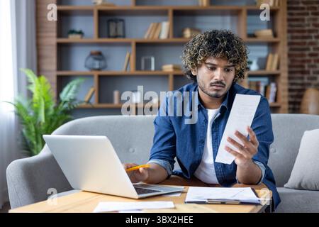 Un uomo con i capelli ricci esamina una ricevuta mentre lavora su un notebook a casa. Foto Stock