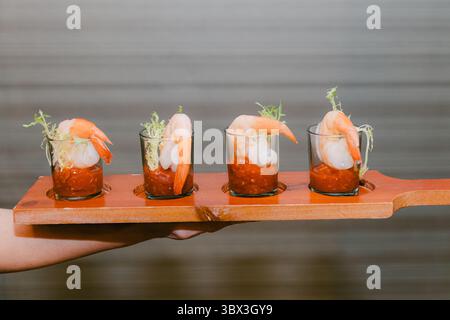 Quattro eleganti cocktail di gamberi sono presentati su un vassoio di legno, guarnito con erbe, pronti per essere gustati. Foto Stock