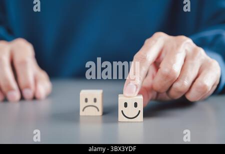 Concetto di soddisfazione del cliente e feedback. Una mano seleziona un blocco di legno con una faccia sorridente su un blocco triste, che rappresenta la soddisfazione del cliente, Foto Stock