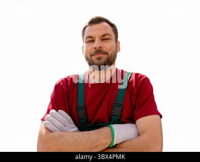 Lavoratore agricolo maschile con barba isolata che indossa tute di bavaglino verde e t-shirt rossa con braccia incrociate che guardano la fotocamera Foto Stock