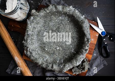 Crosta di torta di sesamo nera ricoperta su un piatto di torta: Impasto salato in una padella con un mattarello, cesoie da cucina e barattolo di farina Foto Stock