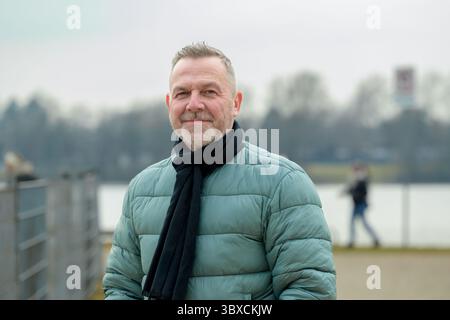Un uomo maturo e sicuro di sé, con una giacca invernale verde e una sciarpa nera, sorride dolcemente sul lago. Foto Stock