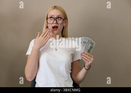 Giovane donna bionda con occhiali e t-shirt bianca dall'espressione scioccata, che regge una grande quantità di dollari USA su un semplice sfondo beige Foto Stock
