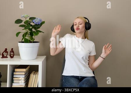 Una donna bionda in un abbigliamento casual e con cuffie wireless nere gode di musica a occhi chiusi e divertenti gesti danzanti mentre è seduta in un ambiente accogliente e moderno Foto Stock