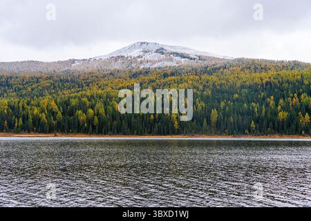 Un lago scuro e calmo con una foresta autunnale verde e gialla lungo il suo litorale, sostenuto da una vetta innevata. Foto Stock