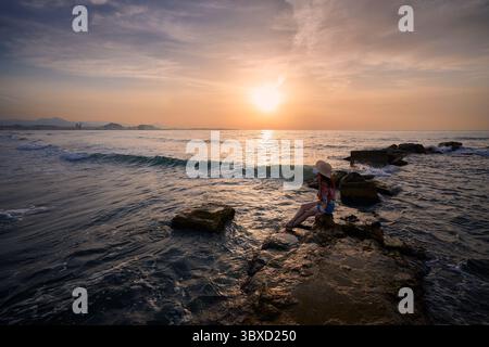 Vista del tramonto sull'oceano con una donna che si rilassa sulla riva Foto Stock