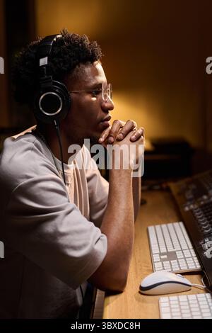 Giovane uomo nero adulto che indossa le cuffie sedute sulla console di mixaggio, concentrandosi sulla produzione audio in studio di registrazione, mani agganciate sotto il mento, profilo laterale visibile Foto Stock