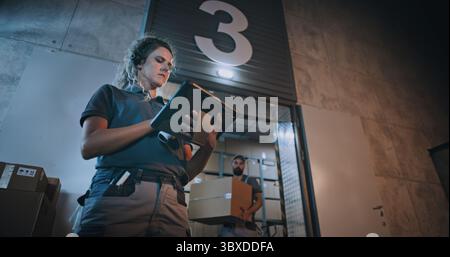 Responsabile donna che esegue la scansione di pacchi con scanner di codici a barre, utilizzando un tablet digitale. Dipendenti diversi che trasportano scatole di cartone dal magazzino di distribuzione logistica di notte. Ordini online, merci, consegna. Foto Stock