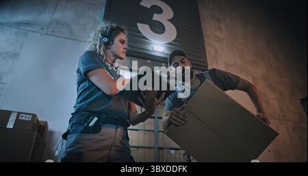 Responsabile donna che esegue la scansione di pacchi con scanner di codici a barre, utilizzando un tablet digitale. Dipendenti diversi che trasportano scatole di cartone dal magazzino di distribuzione logistica di notte. Ordini online, merci, consegna. Foto Stock