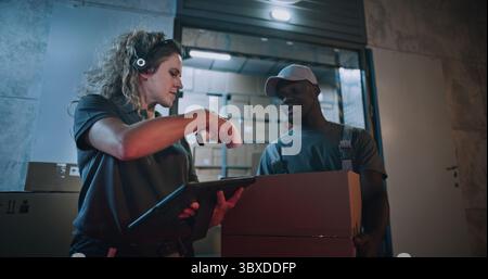 Responsabile dell'inventario femminile che esegue la scansione di scatole di cartone con scanner di codici a barre, utilizzando un computer tablet. Lavoratori multietnici che trasportano pacchi con ordini online, merci dal magazzino di logistica al dettaglio di notte. Foto Stock