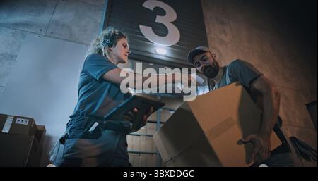 Responsabile donna che esegue la scansione di pacchi con scanner di codici a barre, utilizzando un tablet digitale. Dipendenti diversi che trasportano scatole di cartone dal magazzino di distribuzione logistica di notte. Ordini online, merci, consegna. Foto Stock