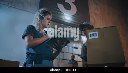 Dipendenti diversi che trasportano scatole di cartone dal magazzino di distribuzione logistica di notte. Responsabile donna che esegue la scansione di pacchi con scanner di codici a barre, utilizzando un tablet digitale. Ordini online, merci, consegna. Foto Stock