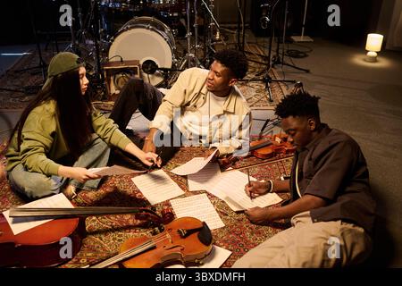 Gruppo multietnico di musicisti che collaborano alla composizione musicale, seduti a terra con violini e spartiti, discutendo idee e scrivendo note in studio di prova Foto Stock