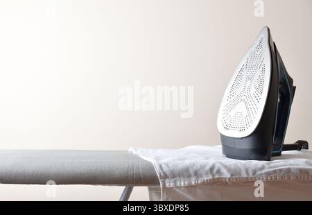 Sistema di stiratura a casa con ferro su asse da stiro grigio con camicia bianca e sfondo isolato chiaro. Vista frontale. Foto Stock