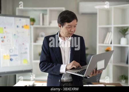 Un anziano esperto finanziario si concentra sul suo notebook in un ufficio elegante, evidenziando il suo ruolo nell'analisi finanziaria e nella pianificazione strategica Foto Stock