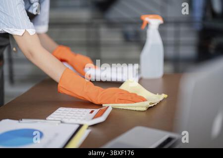 Donna di pulizia per ufficio che indossa guanti arancioni, pulisce una scrivania con materiali e documenti per la pulizia, sottolineando la manutenzione dell'area di lavoro Foto Stock