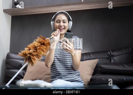 Pulizia della casa con divertimento. Felice giovane casalinga asiatica che canta canzoni durante la pulizia, usando piume come microfono, godendo di lavoro domestico. Giovane Wo Foto Stock