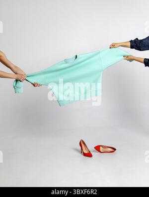 Le mani femminili tengono in mano la felpa color menta e la allungano in diverse direzioni. Scarpe da donna rosse sul pavimento su sfondo grigio. Foto verticale Foto Stock